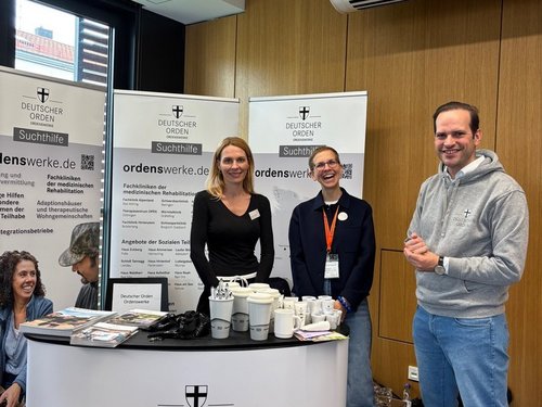 Drei Personen stehen an einem weißen Informationsstand des „Deutschen Ordens“. Im Hintergrund sind drei Banner mit der Webadresse „ordenswerke.de“ zu sehen. Auf dem Tresen liegen Broschüren und stehen Kaffeebecher. Die Aufschrift am Stand lautet „Helfen und Heilen“.