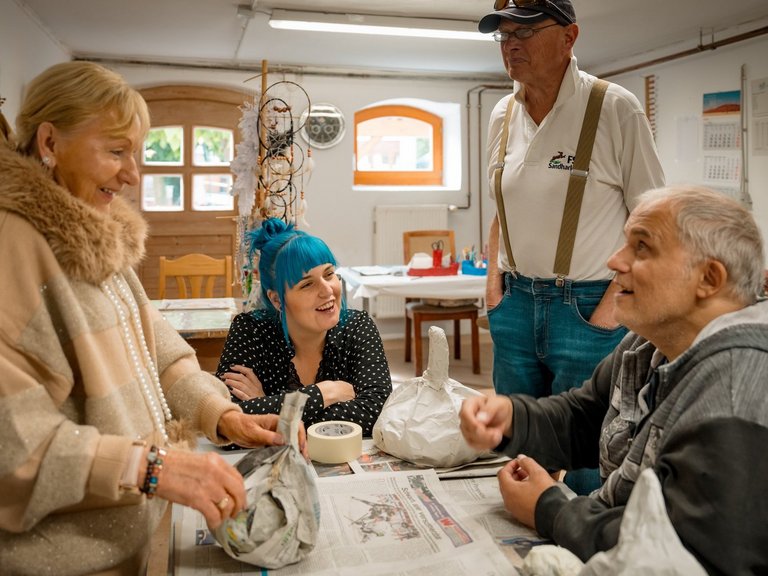 Zwei Frauen und zwei Männer sitzen und stehen an einem Tisch und unterhalten sich, zwei packen Gegenstände in Zeitungspapier.