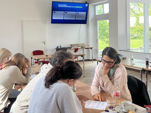 Eine Gruppe von Menschen sitzt an einem Tisch in einem hellen Raum und bespricht Dokumente. Im Hintergrund zeigt ein Bildschirm eine Präsentation mit dem Titel „Workshop“. Auf dem Tisch stehen Getränke und Snacks.