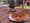 Eine Pizza auf einem Papierteller auf einem Holztisch im Freien. Im Hintergrund sind ein belebter Markt, ein orangenes Zelt und Spaziergänger bei sonnigem Wetter zu sehen.